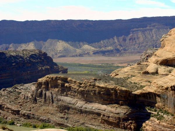 A panoramic view of rugged rock formations and cliffs in a canyon landscape, with layered mountains in the background and a valley visible below. The scene showcases earthy tones and textures of the rock, under a partly cloudy sky. Slickrock mountain bike trail.