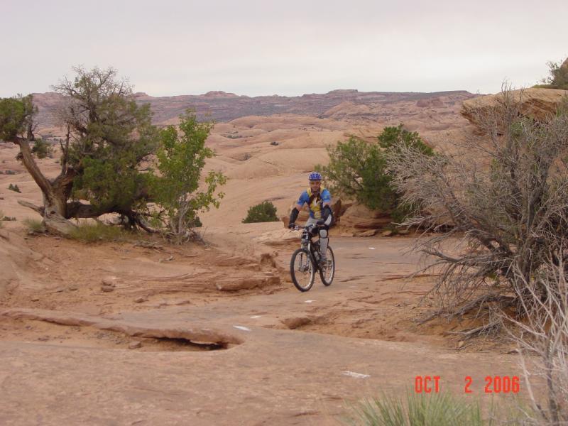 A mountain biker rides along a rugged dirt trail in a desert landscape, surrounded by rocky terrain and sparse vegetation. The sky is overcast, hinting at a cool atmosphere, while the diverse textures of the ground and plants create a natural setting for outdoor adventure. Slickrock mountain bike trail.