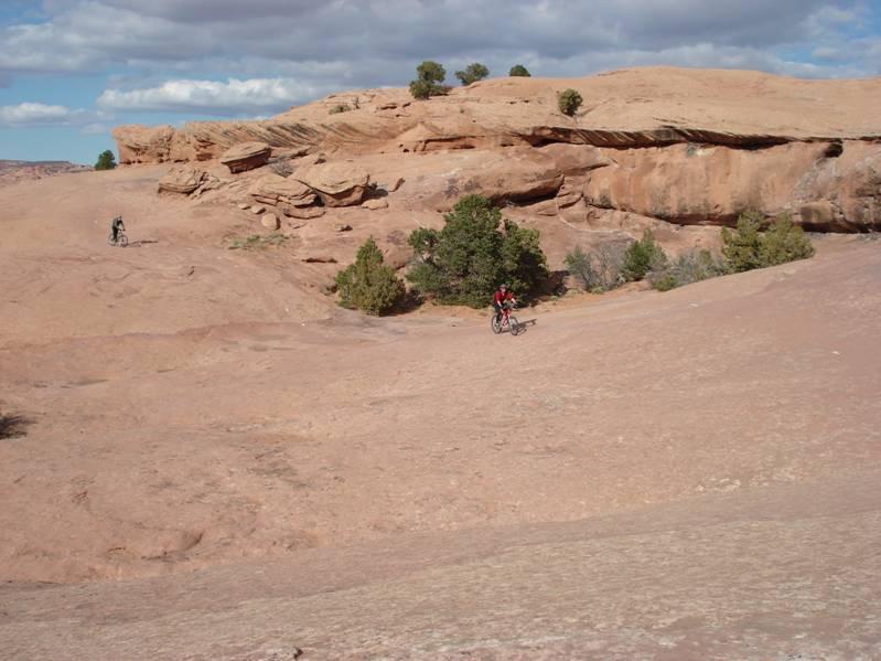 Two mountain bikers traverse a rugged, rocky landscape under a cloudy sky. The terrain features distinct rock formations and sparse vegetation, with scattered bushes dotting the scene. The setting suggests a remote outdoor adventure location. Slickrock mountain bike trail.