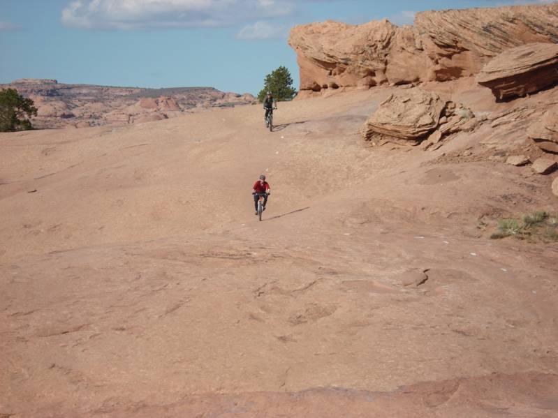 Two mountain bikers riding on a rocky, desert landscape with red stone formations and a clear blue sky. One biker is near the foreground, while the other is further up the slope. Sparse vegetation is visible among the rocks. Slickrock mountain bike trail.