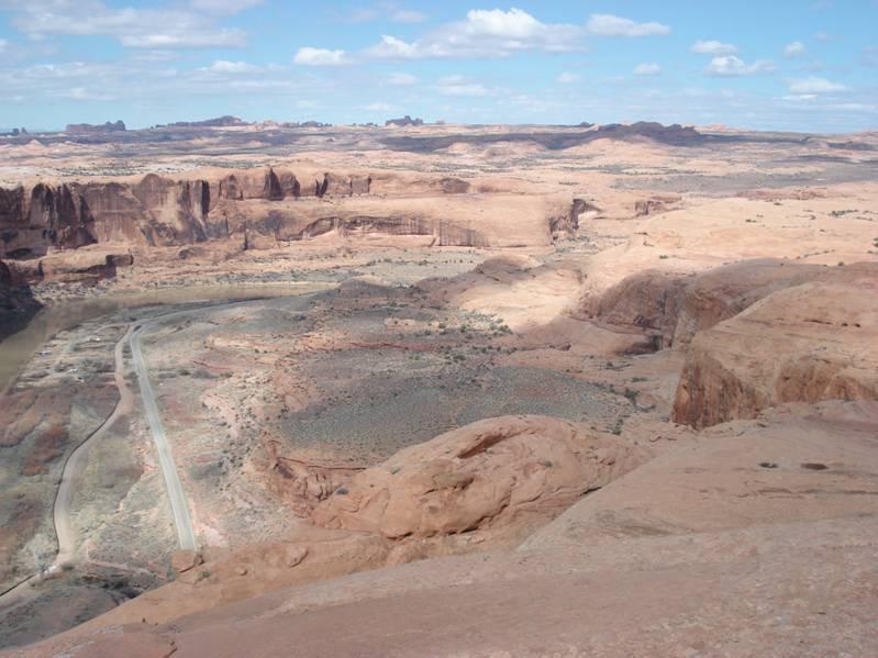 A panoramic view of a desert landscape featuring rocky formations, gentle slopes, and a winding road along a river. The sky is partly cloudy, and the scene showcases the vastness of the arid terrain with patches of greenery scattered throughout. Slickrock mountain bike trail.