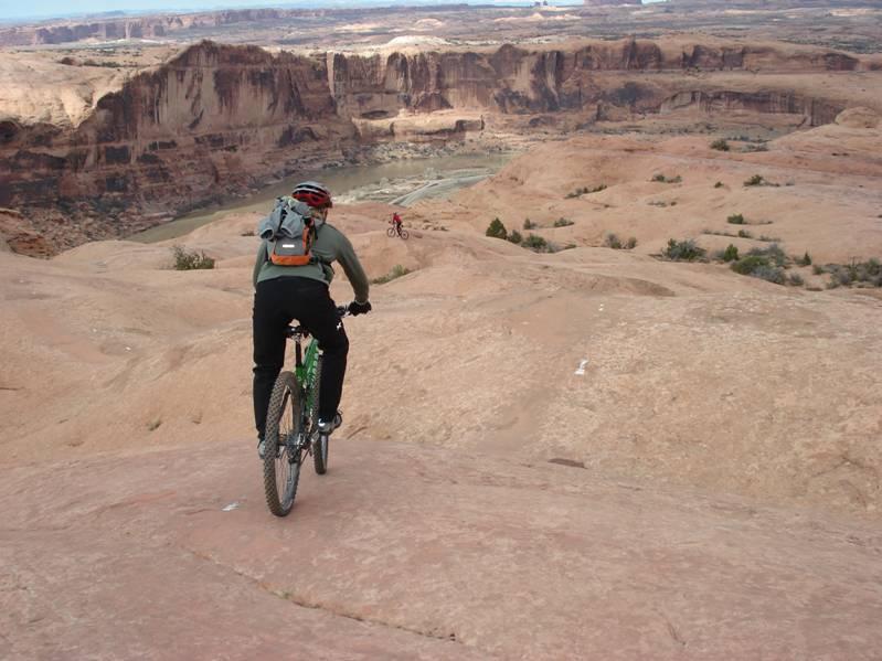 A mountain biker descending a rocky ridge with a downhill path that leads to a canyon below. The landscape features red rock formations and a river winding through the canyon in the distance. Another cyclist can be seen in the background, demonstrating the adventurous nature of the terrain. Slickrock mountain bike trail.