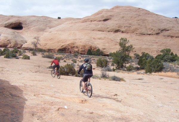 Two mountain bikers are riding on a rocky, arid terrain, surrounded by sparse vegetation and large, sandy rock formations. The sky is cloudy, and the landscape features a mix of shrubs and small trees. Slickrock mountain bike trail.