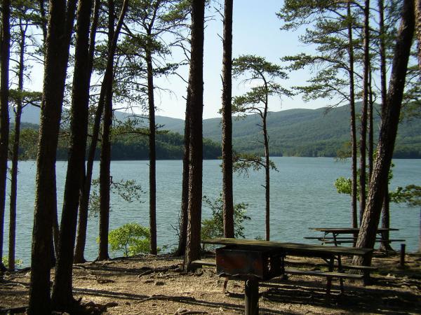A serene view of a lake surrounded by tall trees, with a picnic table in the foreground. The sun shines brightly over the water, reflecting the mountains in the background and creating a peaceful outdoor atmosphere. Carvin's Cove Trail system mountain bike trail.