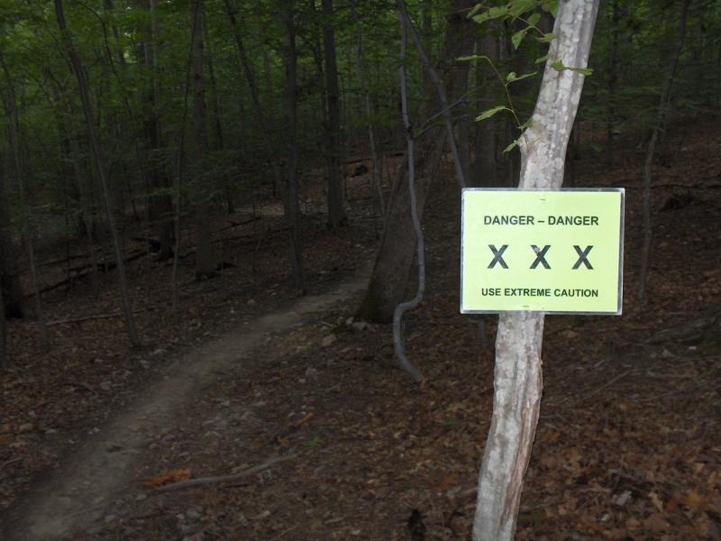Sign warning of danger and advising extreme caution, displayed on a tree in a wooded area with a dirt path visible in the background. Fountainhead Regional Park mountain bike trail.
