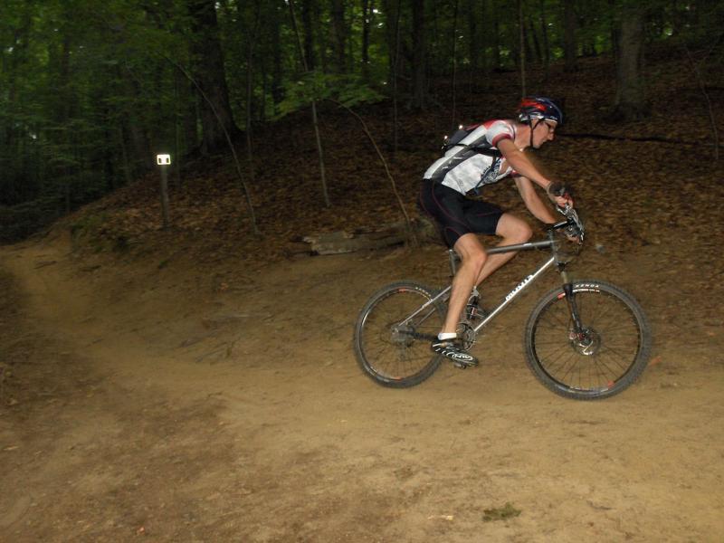 A mountain biker in a cycling jersey and helmet navigating a dirt trail surrounded by trees. The biker is mid-action, leaning forward as they ride over a curve in the path. The forest floor is covered with fallen leaves, and a trail marker is visible in the background. Fountainhead Regional Park mountain bike trail.