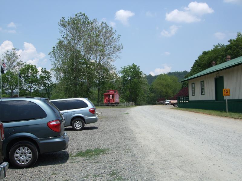 A gravel parking area with several parked vehicles, surrounded by trees and blue skies with scattered clouds. In the background, a red train caboose is visible next to a green building. A sign indicating a speed limit of 15 mph is also present. New River Trail State Park mountain bike trail.