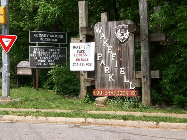A wooden sign for Wakefield Park displayed prominently next to a road, with information about the park and an adjacent sign for the Audrey Moore Rec Center. The sign includes details about a spring and summer membership sale, along with a phone number for more information. The background features green foliage. Wakefield mountain bike trail.