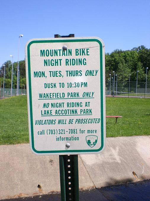 Signage for mountain bike night riding regulations, detailing allowed days (Monday, Tuesday, Thursday), hours (dusk to 10:30 PM) at Wakefield Park only, with a prohibition against night riding at Lake Accotink Park and a warning that violators will be prosecuted. Contact information is provided for further inquiries. Wakefield mountain bike trail.