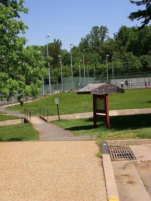 A sunny park scene featuring a grassy area with a pathway leading to a tennis court. The background includes several tennis courts with players in action, surrounded by trees and clear blue skies. A wooden information kiosk stands on the path, providing details about the park. Wakefield mountain bike trail.