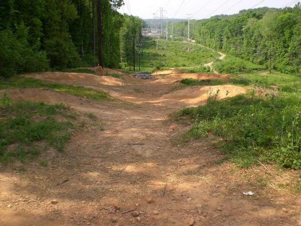 A dirt path leading downhill through a wooded area, with grassy edges and a few small bumps along the track. Power lines are visible in the background, and a clearing can be seen in the distance. The scene is surrounded by trees and natural vegetation, suggesting a recreational area for biking or hiking. Wakefield mountain bike trail.