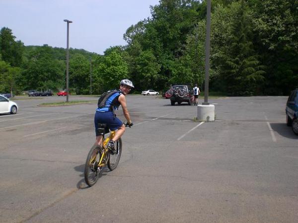 A cyclist wearing a helmet and a blue jersey rides a mountain bike in a parking lot surrounded by trees. Several cars are parked in the background, and another person is seen loading a bike onto a vehicle. Wakefield mountain bike trail.