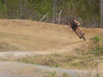 A person in a black tank top and helmet riding a mountain bike on a dirt track, leaning into a turn surrounded by greenery and trees. Wakefield mountain bike trail.