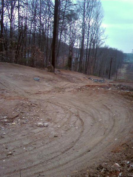 A winding dirt path surrounded by bare trees, with a cloudy sky overhead. The ground is uneven, showing signs of recent disturbance or construction, and there are rocks scattered along the track. Wakefield mountain bike trail.