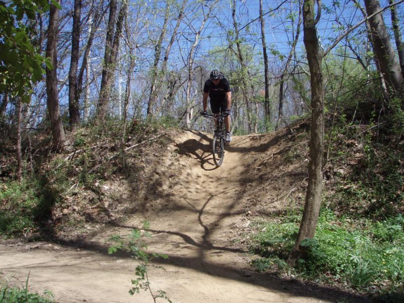 A person riding a mountain bike down a dirt path with a slope in a wooded area on a sunny day. The scene is surrounded by trees and greenery, showcasing a vibrant outdoor environment. Wakefield mountain bike trail.