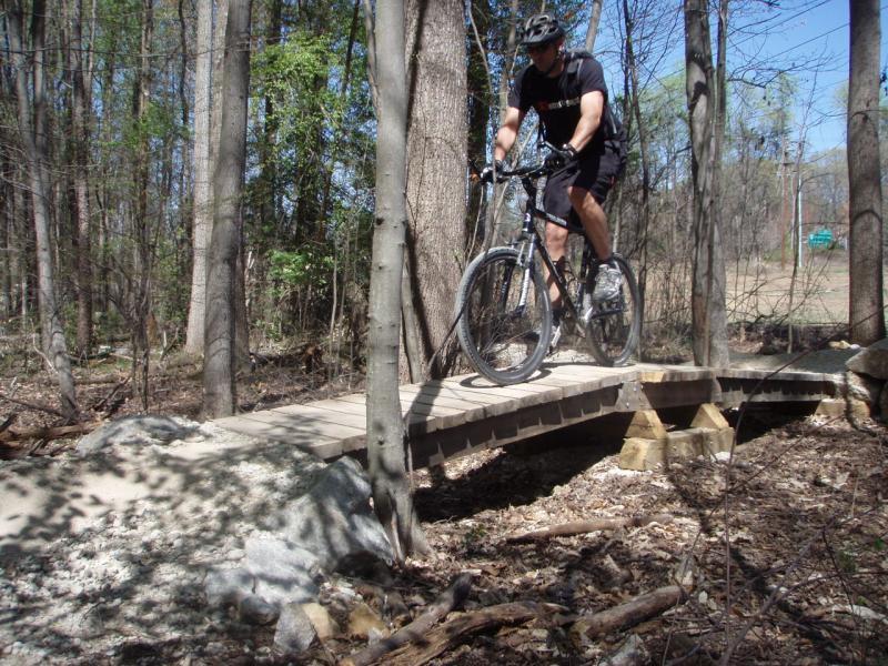 A person riding a mountain bike crosses a wooden bridge in a wooded area, surrounded by trees and natural foliage. The scene captures a sunny day with clear skies and a mix of dirt and gravel on the ground. Wakefield mountain bike trail.