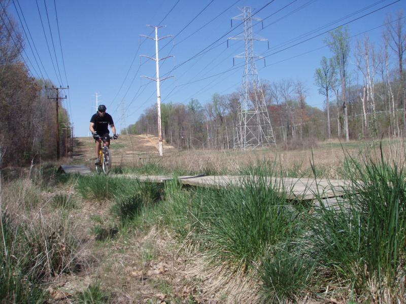 A mountain biker riding on a wooden path through grassy terrain, with power lines and trees in the background on a clear day. Wakefield mountain bike trail.