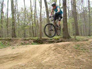 A mountain biker in mid-air jumps over a small dirt ramp in a forested area, surrounded by trees and greenery. The rider is wearing a blue tank top, black shorts, and a helmet, demonstrating an action-packed moment of biking. Wakefield mountain bike trail.