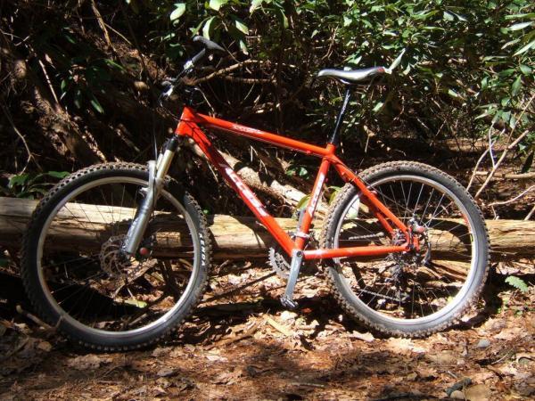 A bright orange mountain bike resting on the ground among fallen leaves and branches in a wooded area, with greenery in the background. The bike features knobby tires and a sturdy frame suitable for off-road riding. Bear Creek mountain bike trail.
