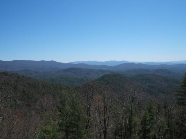 A panoramic view of rolling mountains under a clear blue sky, with layers of green and brown foliage in the foreground and distant peaks fading into the horizon. Bear Creek mountain bike trail.