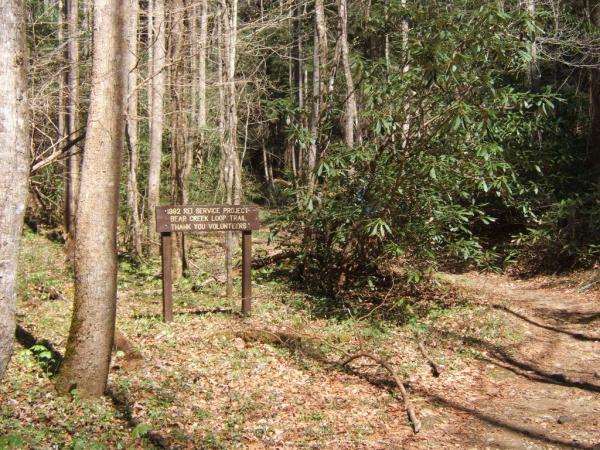 A wooden sign in a wooded area reads "2002 REI SERVICE PROJECT BEAR DEN LOOP TRAIL THANK YOU VOLUNTEERS," with trees and shrubs surrounding the trail entrance. Bear Creek mountain bike trail.