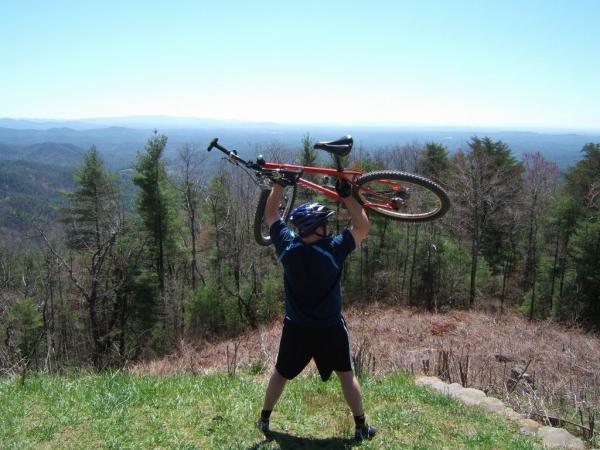 A person wearing a helmet and athletic clothing stands on a hillside, lifting a mountain bike above their head. In the background, a vast landscape of rolling hills and trees stretches out under a clear blue sky. Bear Creek mountain bike trail.