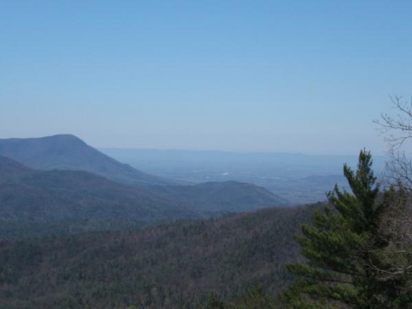 A panoramic view of a mountainous landscape under a clear blue sky, featuring rolling hills and dense forests. The scene captures the vastness of the terrain with gentle slopes leading to distant peaks, showcasing natural beauty and tranquility. Bear Creek mountain bike trail.