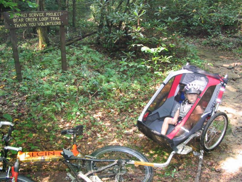 A child in a bike trailer sits on a forest path next to a mountain bike, with a sign that reads "REI Service Project Bear Creek Loop Trail Thank You Volunteers" in the background, surrounded by lush greenery. Bear Creek mountain bike trail.