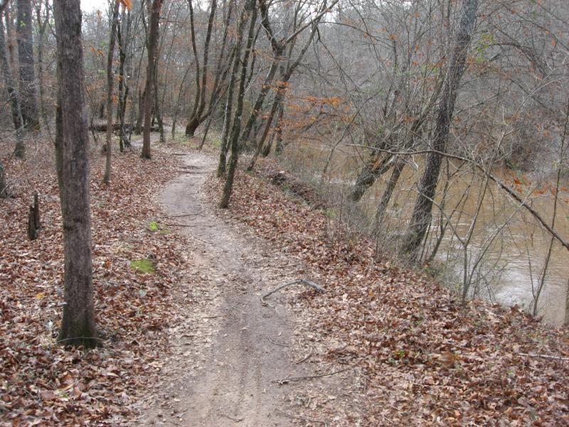 A winding dirt path surrounded by bare trees and fallen leaves, leading alongside a river in a wooded area. The landscape appears to be in a late autumn or early winter setting, with a muted color palette. Heritage Park mountain bike trail.