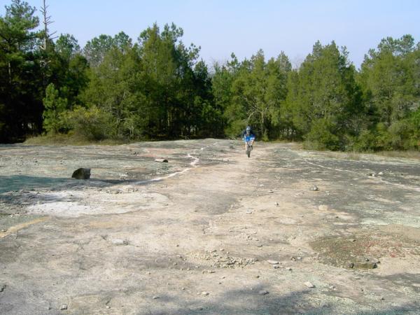 A person walking on a rocky terrain surrounded by trees, with a clear sky in the background. The landscape features a mostly flat surface with scattered stones and patches of earth. Georgia International Horse Park mountain bike trail.