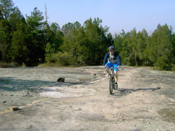 A mountain biker dressed in blue shorts and a long-sleeve shirt rides along a rocky trail surrounded by green trees under a clear sky. Georgia International Horse Park mountain bike trail.