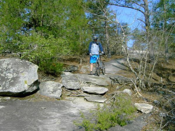 A cyclist standing beside a mountain bike on a rocky trail surrounded by trees and greenery. The trail appears rugged, with large stones and a clear blue sky in the background. The cyclist is wearing a helmet and athletic clothing. Georgia International Horse Park mountain bike trail.