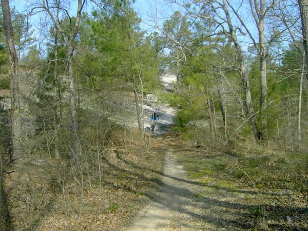A scenic dirt path winding through a wooded area, surrounded by trees. In the distance, a person is walking along the trail, with blue skies visible above. The setting suggests a peaceful outdoor environment ideal for hiking and exploring nature. Georgia International Horse Park mountain bike trail.