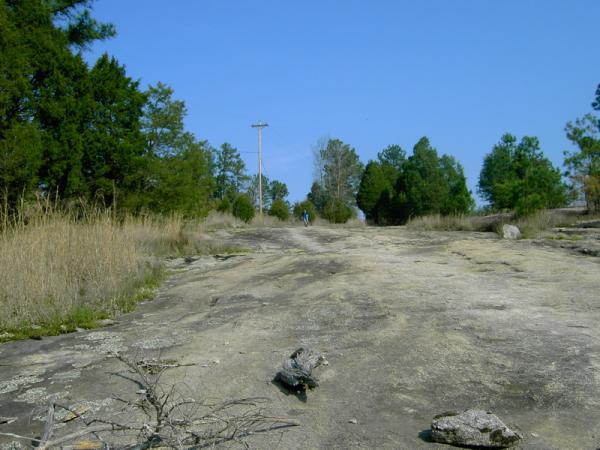 A rocky path leads through a grassy area, bordered by trees on either side. In the distance, a person walks along the path under a clear blue sky, while a power line runs nearby. The scene is serene, showcasing natural elements and open space. Georgia International Horse Park mountain bike trail.