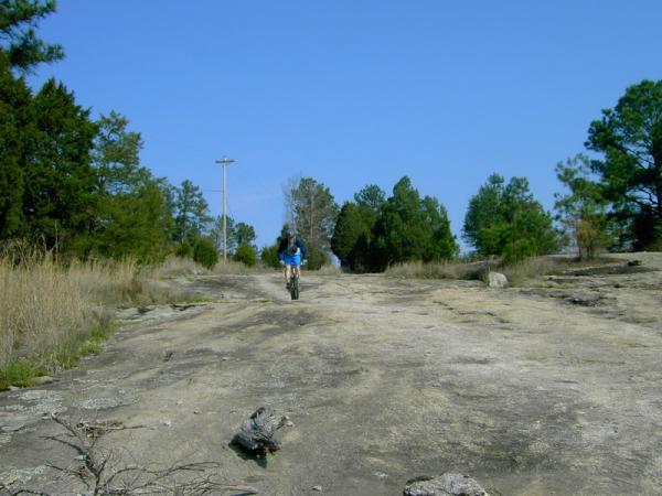 A person riding a bicycle along a rocky, uneven path surrounded by trees and a clear blue sky. The landscape includes patches of grass and small boulders, with a power line visible in the background. Georgia International Horse Park mountain bike trail.