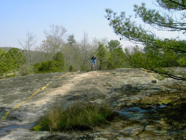 A person walking on a large, smooth rock surface surrounded by sparse vegetation and trees under a clear blue sky. Yellow markings are visible on the rock, guiding the path. Georgia International Horse Park mountain bike trail.