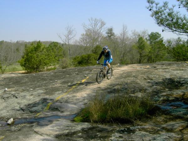 A mountain biker wearing a helmet and cycling gear rides along a rocky trail marked by a yellow line, surrounded by sparse trees and a clear blue sky. Georgia International Horse Park mountain bike trail.