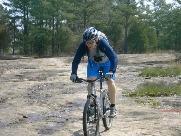 A person wearing a helmet and cycling gear rides a mountain bike over a rocky terrain, surrounded by trees in the background. The cyclist appears focused and is navigating the uneven surface. Georgia International Horse Park mountain bike trail.