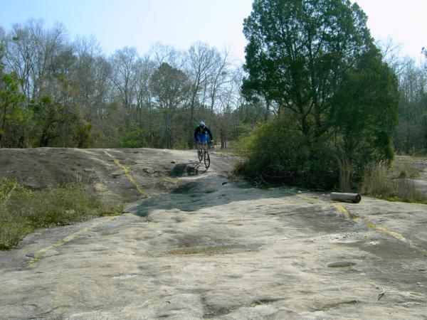 A mountain biker riding on a rocky, uneven trail surrounded by sparse trees and brush. The landscape is mostly bare rock with faint yellow lines marking the path. The sky is clear and blue, indicating a sunny day. Georgia International Horse Park mountain bike trail.