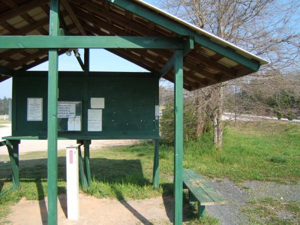 A green wooden shelter with a sloped roof, featuring a bulletin board displaying various notices. To the right, there is a bench under the shelter, surrounded by grass and trees. A dirt path leads away from the shelter towards a nearby road. The sky is clear and sunny. Georgia International Horse Park mountain bike trail.