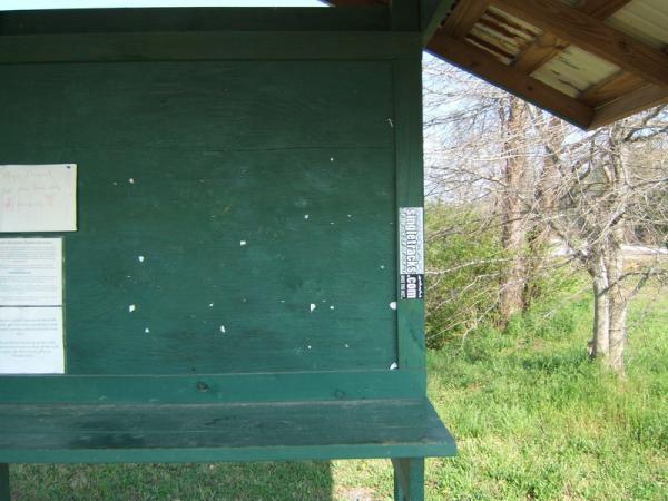 A green bulletin board located in a rural area, featuring a few pieces of paper attached to its surface. The board is mostly empty, with some small holes visible. In the background, there are trees and open grass, indicating a natural setting. Georgia International Horse Park mountain bike trail.