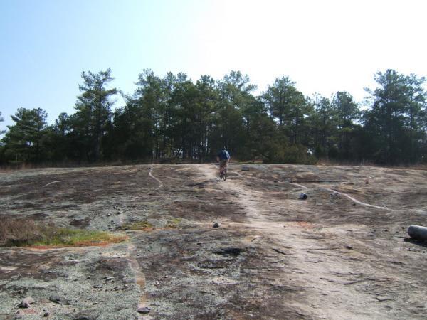 A lone cyclist ascending a rocky, sloped area with patches of moss and lichen, surrounded by a dense tree line in the background under a clear blue sky. Georgia International Horse Park mountain bike trail.
