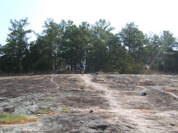 A person walking along a rocky, barren landscape, with a trail leading through a sunny area and a backdrop of tall pine trees. Georgia International Horse Park mountain bike trail.