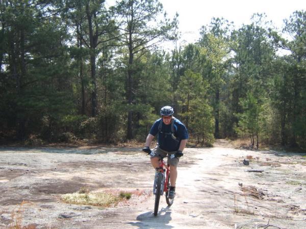 A person riding a mountain bike on a rocky path in a wooded area, surrounded by tall trees and greenery. The cyclist is wearing a helmet and casual athletic clothing, enjoying the outdoors. Georgia International Horse Park mountain bike trail.
