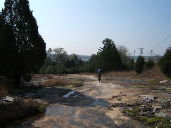 A person walking along a rocky, uneven terrain surrounded by sparse trees, with a clear sky above and distant power lines visible in the background. The landscape features patches of dry grass and scattered debris. Georgia International Horse Park mountain bike trail.