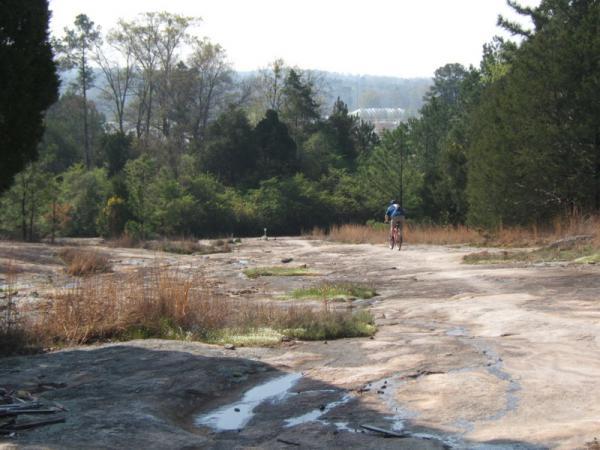 A person riding a mountain bike on a rocky trail surrounded by sparse vegetation and trees, with a distant view of hills and a clear sky in the background. Georgia International Horse Park mountain bike trail.