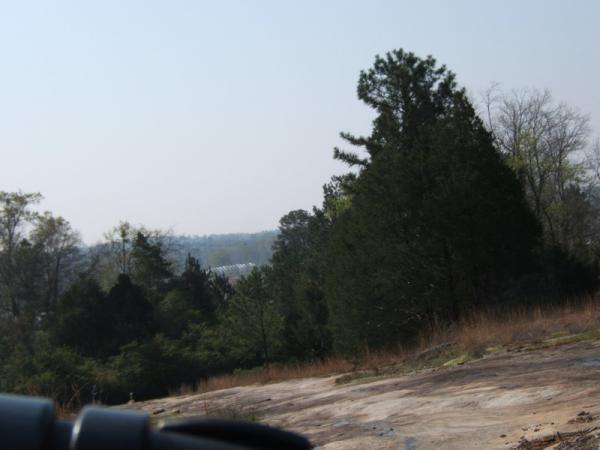 A view of a rocky terrain with patches of grass and various evergreen trees in the foreground. In the background, a misty landscape can be seen with more trees and a glimpse of structures partially obscured by fog. The image captures a serene, natural setting. Georgia International Horse Park mountain bike trail.