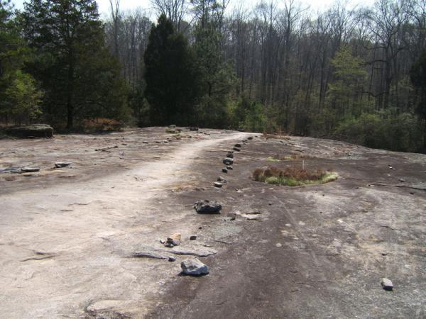 A rocky, flat landscape with a winding path made of stones, surrounded by sparse vegetation and trees in the background. The scene shows a natural area with remnants of rocks scattered across the surface, hinting at a tranquil outdoor setting. Georgia International Horse Park mountain bike trail.