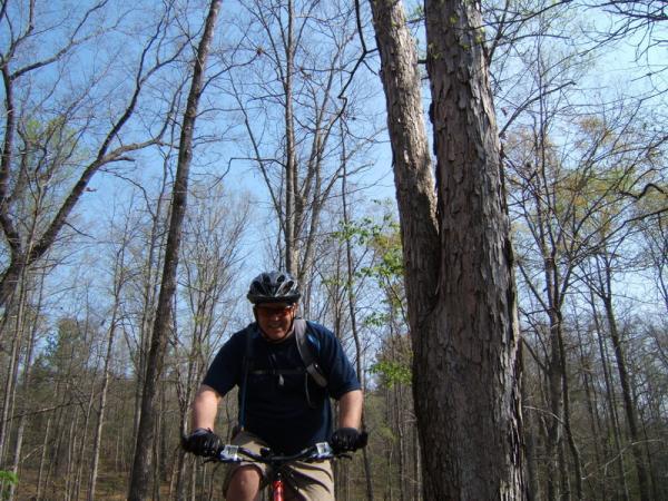 A mountain biker riding along a wooded trail with bare trees in the background under a clear blue sky. The cyclist is wearing a helmet and gloves, focused on the path ahead. Georgia International Horse Park mountain bike trail.