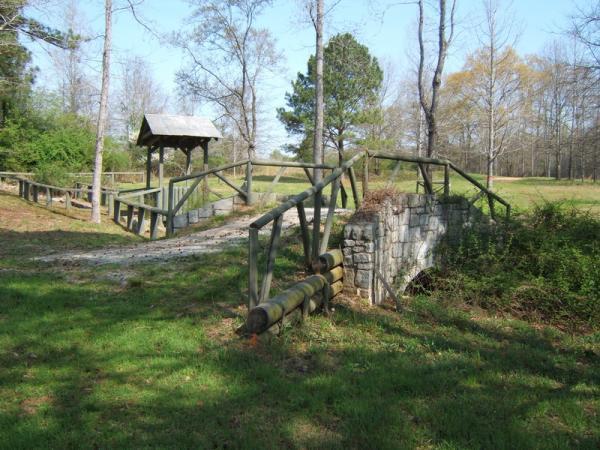 A wooden bridge with railings crosses a small stream in a grassy area, surrounded by trees. A small shelter is visible near the bridge, and a stone structure can be seen to the right. The scene is peaceful, showcasing natural greenery and a clear sky. Georgia International Horse Park mountain bike trail.